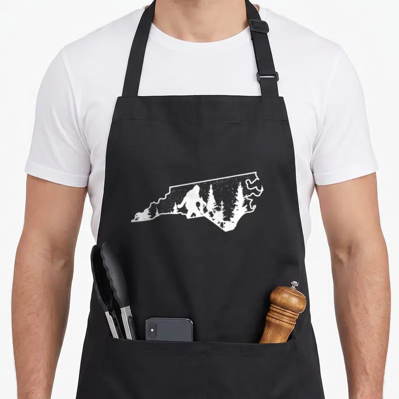 North bigfoot apron - White outline of North Carolina state map containing Bigfoot silhouette and pine trees — shown on a black cotton canvas apron against a white background
