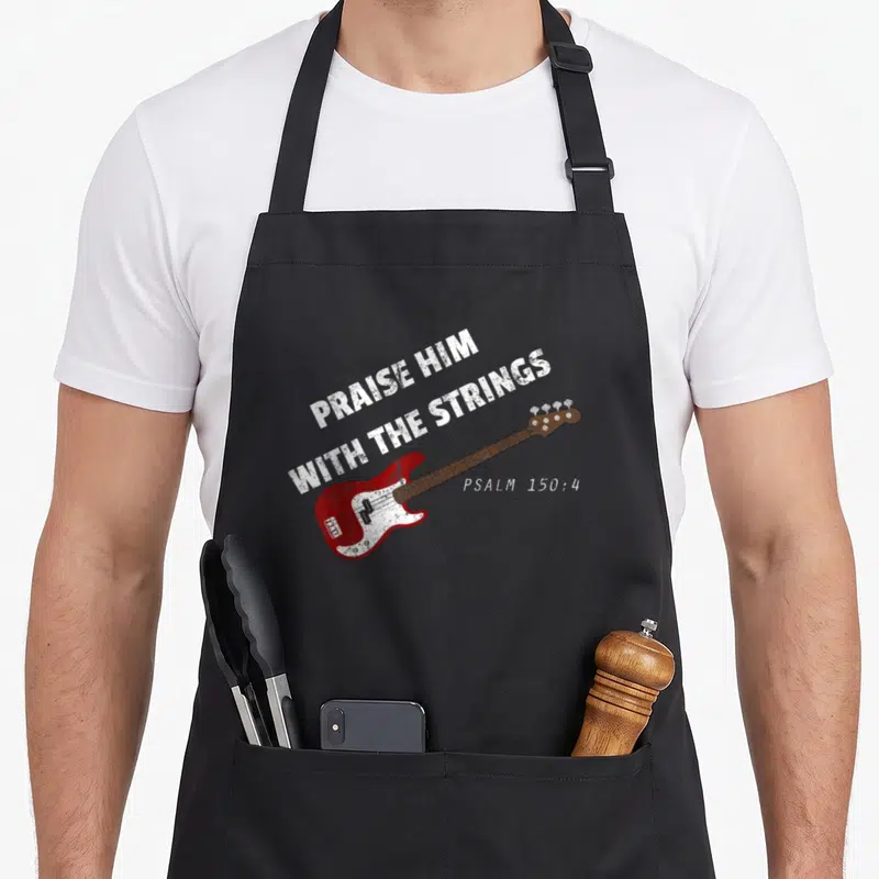 Guitar guitarist christian apron - Distressed white text Praise Him With The Strings above a red bass guitar and Psalm — shown on a black cotton canvas apron against a white background