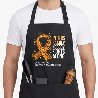 Adhd family aprons - Orange heart awareness ribbon and In This Family Nobody Fights Alone text — shown on a black cotton canvas apron against a white background