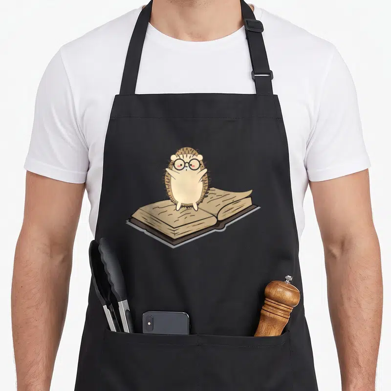 Animal nerd apron - Cartoon hedgehog with glasses standing behind an open book in beige and brown tones — shown on a black cotton canvas apron against a white background