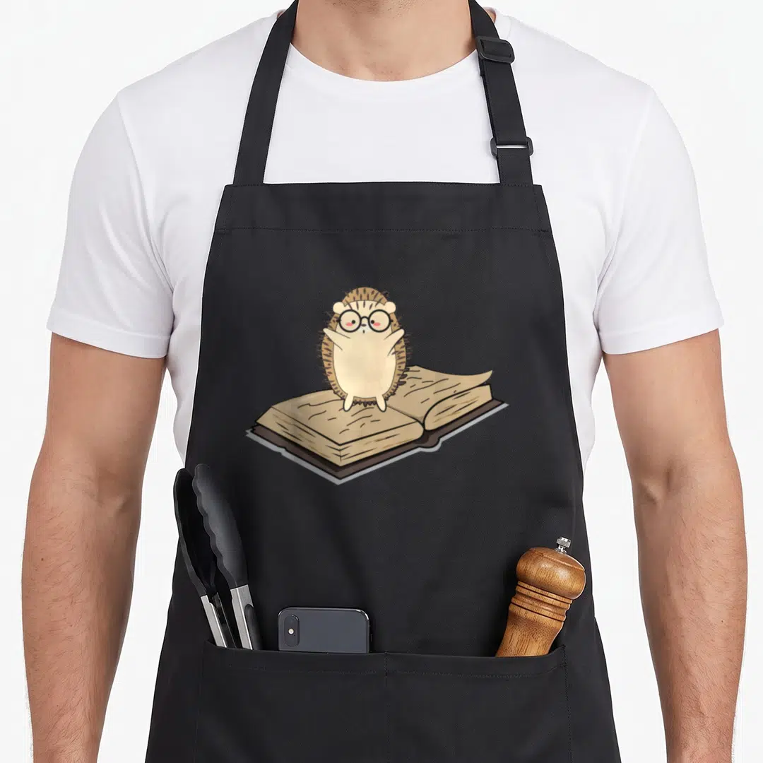Animal nerd apron - Cartoon hedgehog with glasses standing behind an open book in beige and brown tones — shown on a black cotton canvas apron against a white background