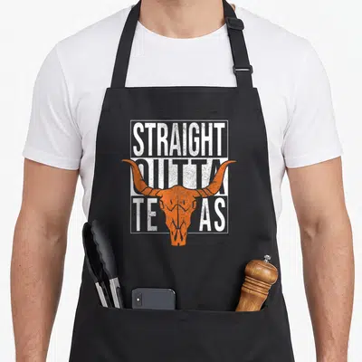 Straight texans apron - Orange bull skull over distressed white text STRAIGHT OUTTA TEXAS in rectangular frame — shown on a black cotton canvas apron against a white background
