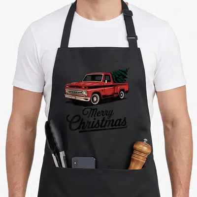 Pickup christmas tree apron - Red vintage truck carrying green Christmas tree above Merry Christmas script in retro — shown on a black cotton canvas apron against a white background