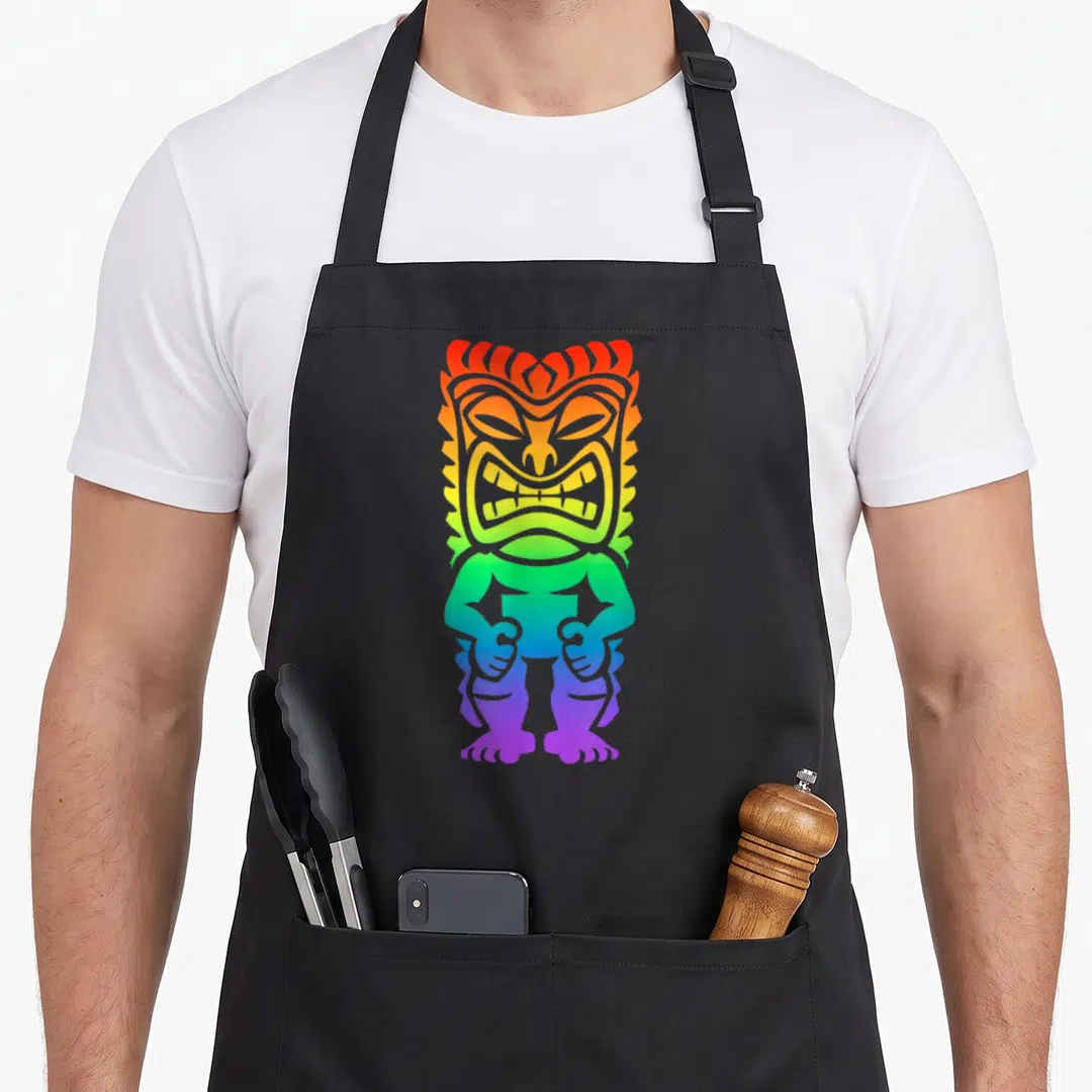 Tiki gay pride apron - Rainbow gradient cartoon tiki statue on black background — shown on a black cotton canvas apron against a white background
