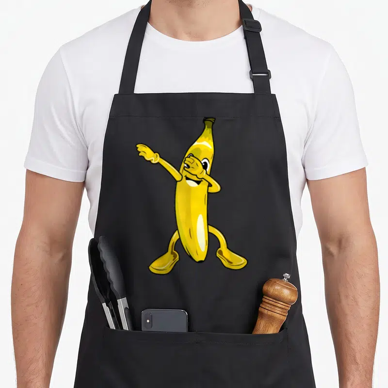 Dab banana apron - Yellow cartoon banana character with one hand covering eye and arm extended to side — shown on a black cotton canvas apron against a white background