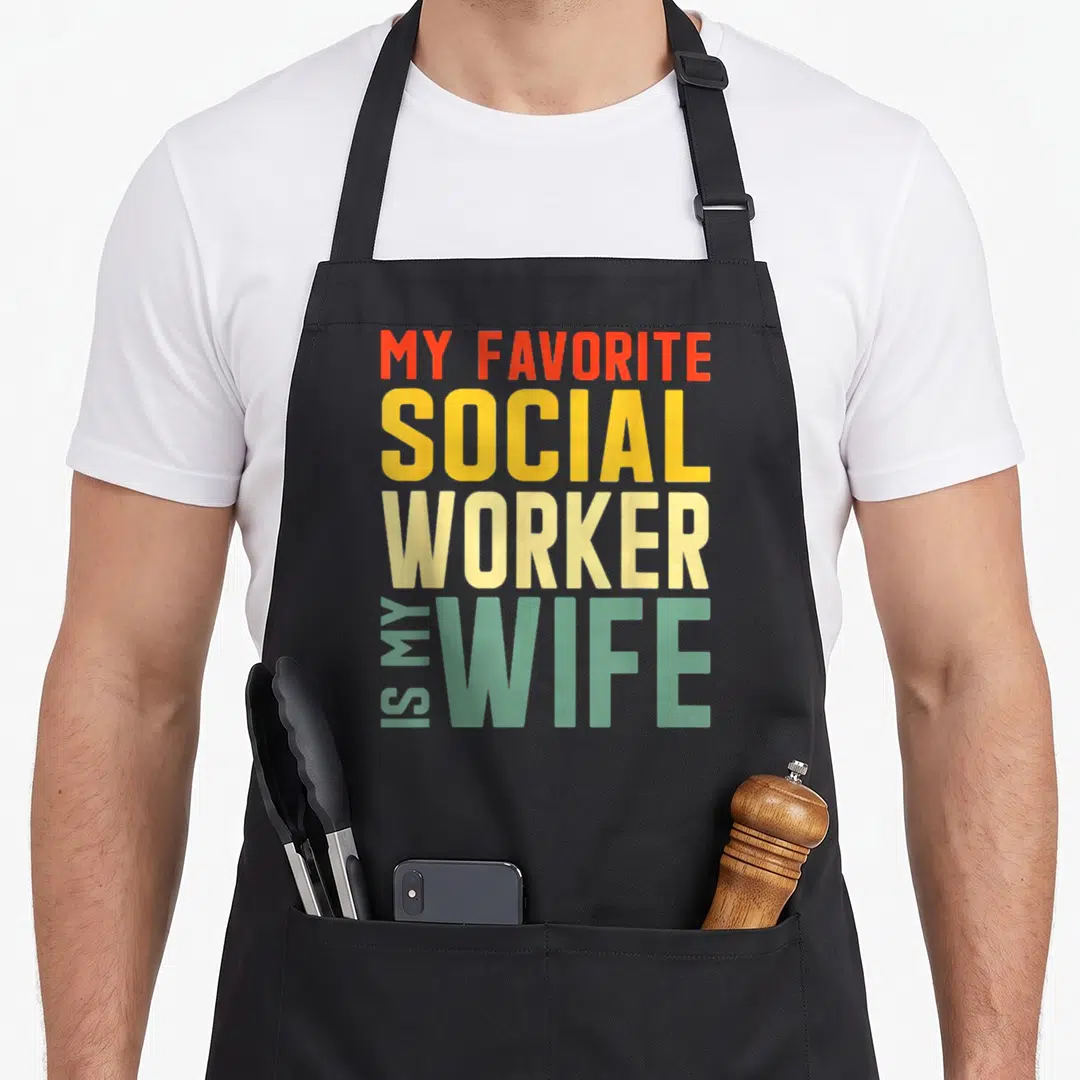 Favourite wife apron - Stacked text MY FAVORITE SOCIAL WORKER IS MY WIFE in orange yellow beige and light green — shown on a black cotton canvas apron against a white background