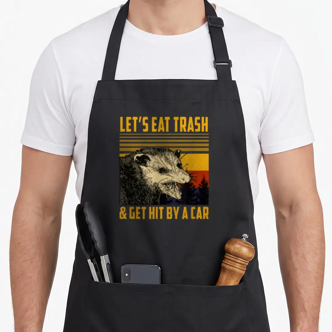 Eat car apron - Angry opossum head over striped sunset with LET'S EAT TRASH text — shown on a black cotton canvas apron against a white background