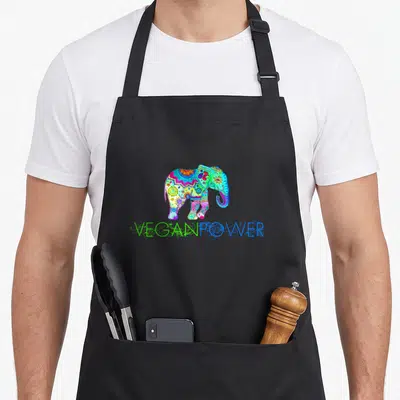 Power vegan apron - Multicolored cartoon elephant with mandala patterns above VEGANPOWER text — shown on a black cotton canvas apron against a white background