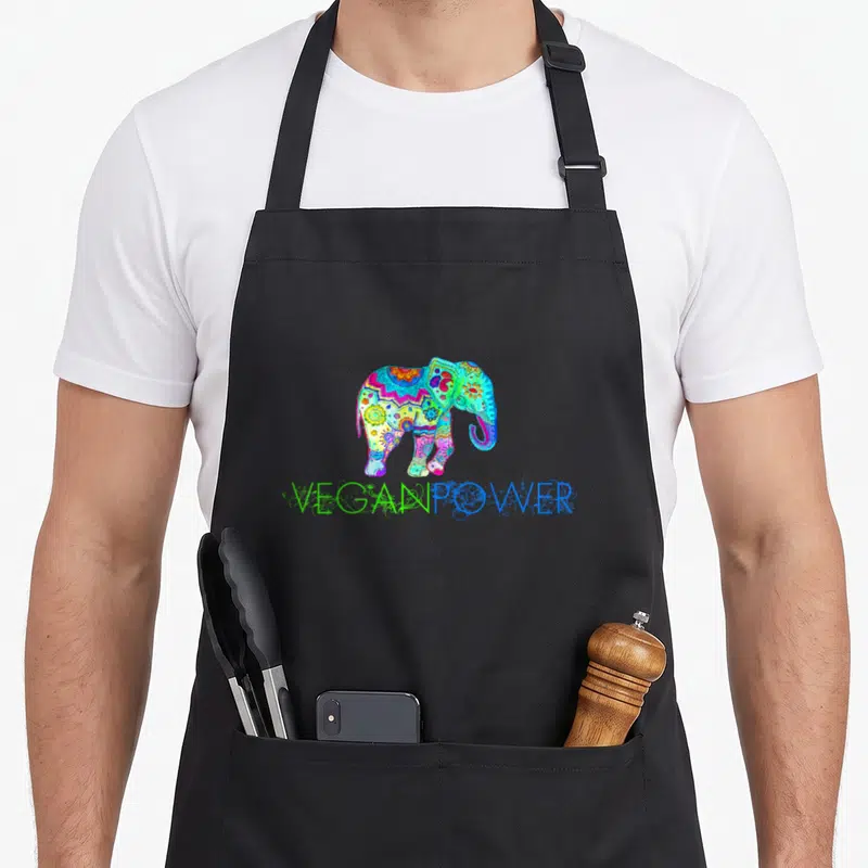 Power vegan apron - Multicolored cartoon elephant with mandala patterns above VEGANPOWER text — shown on a black cotton canvas apron against a white background