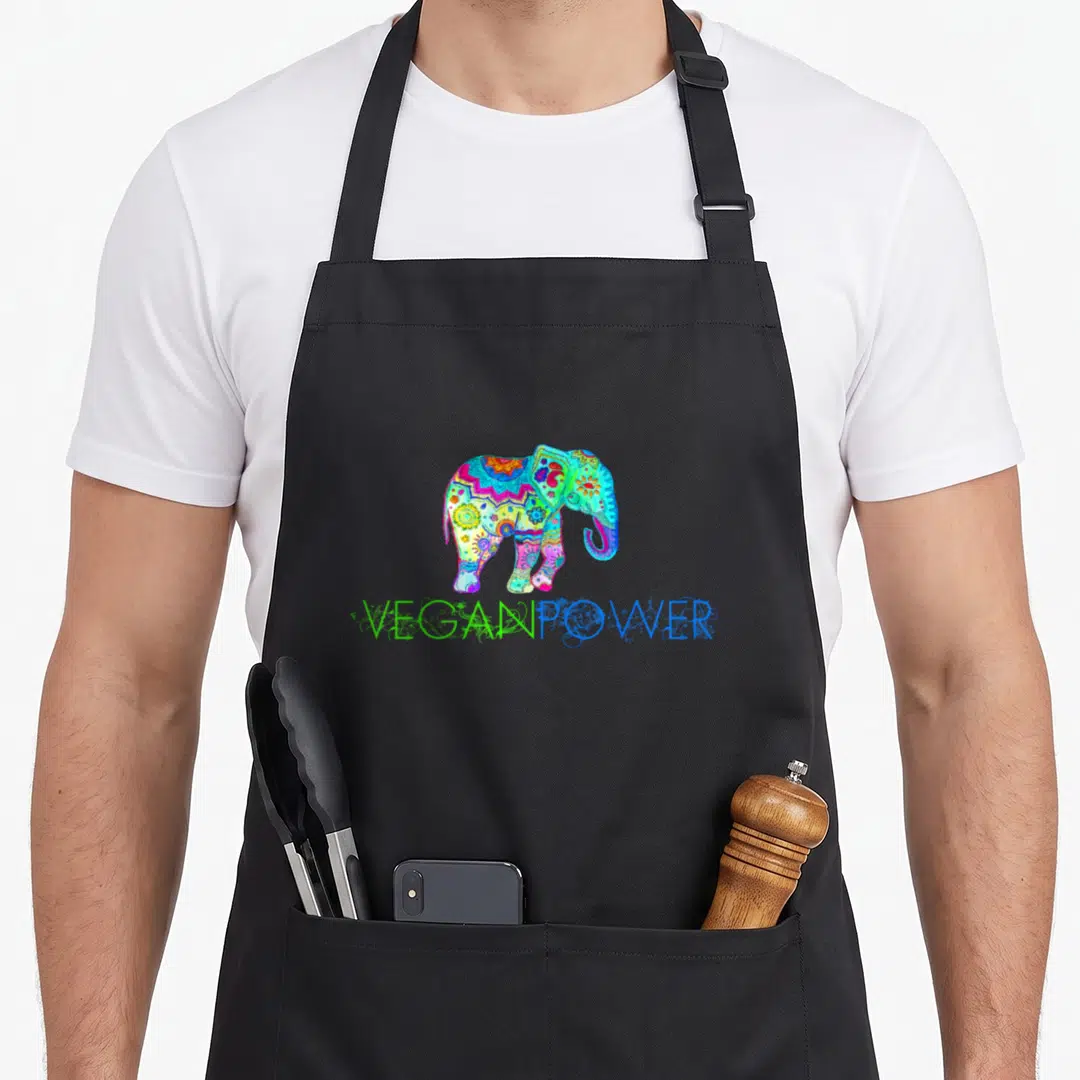 Power vegan apron - Multicolored cartoon elephant with mandala patterns above VEGANPOWER text — shown on a black cotton canvas apron against a white background