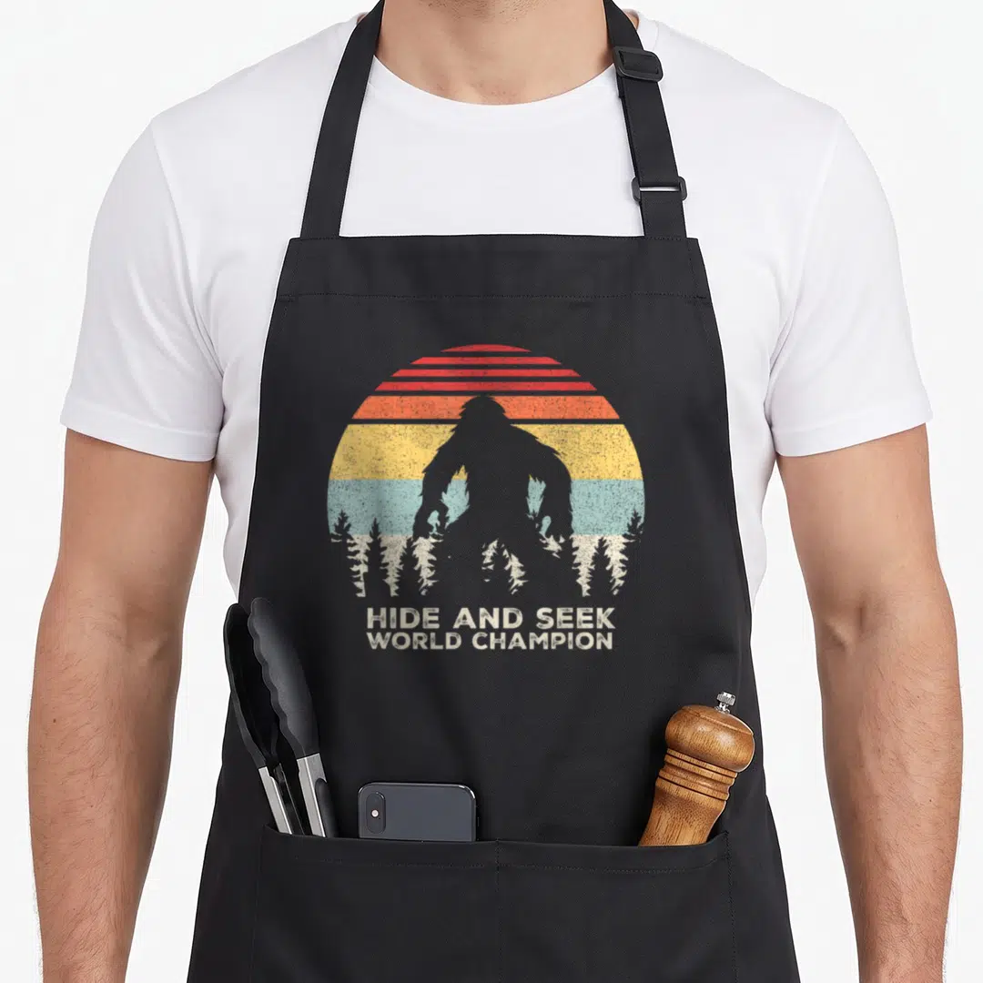 Hide seek champion sasquatch bigfoot apron - Retro bigfoot silhouette against striped sun with pine trees and hide — shown on a black cotton canvas apron against a white background