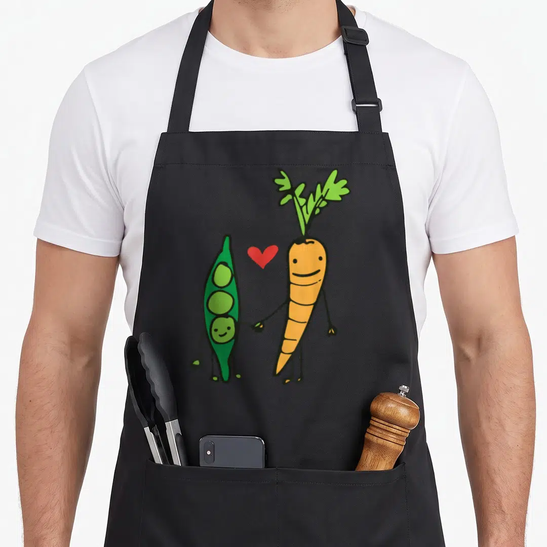 Together family aprons - Cartoon orange carrot and green pea pod holding hands with a red heart between them — shown on a black cotton canvas apron against a white background