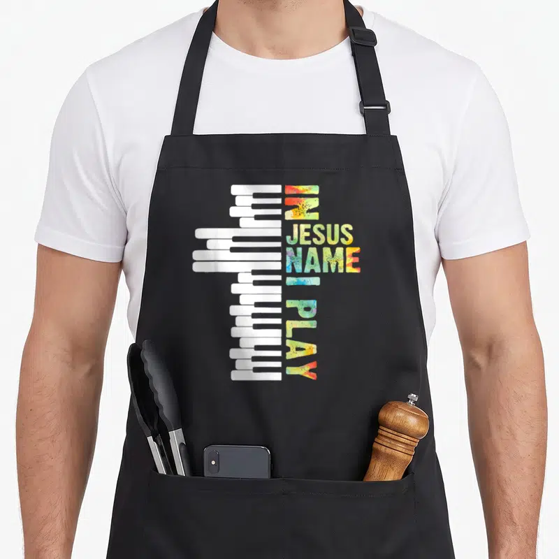 Piano christian apron - Piano keys forming a cross beside vertical IN JESUS NAME I PLAY text in watercolor colors — shown on a black cotton canvas apron against a white background