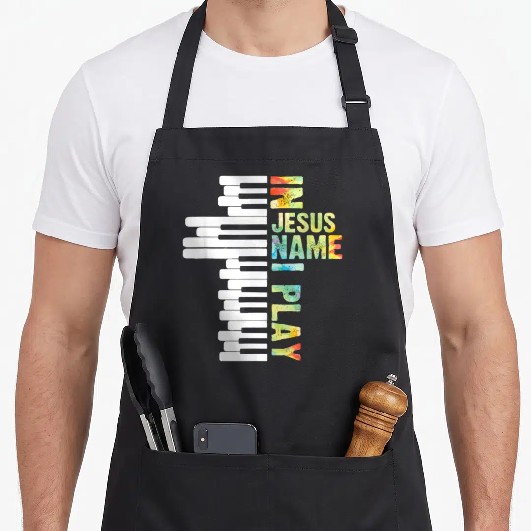 Piano christian apron - Piano keys forming a cross beside vertical IN JESUS NAME I PLAY text in watercolor colors — shown on a black cotton canvas apron against a white background