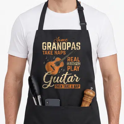 Grandpas acoustics apron - Orange guitar graphic with text about grandpas playing guitar and taking naps in retro style — shown on a black cotton canvas apron against a white background