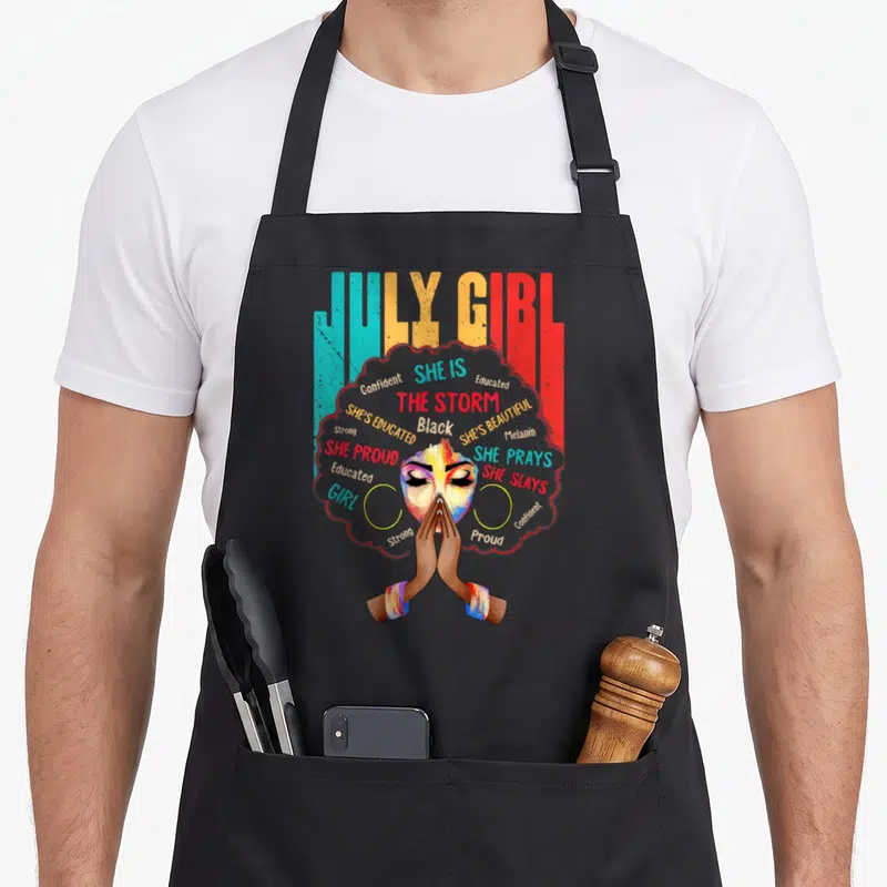Cute july birthday apron - Watercolor woman face with afro made of empowering words like SHE IS THE STORM and JULY GIRL — shown on a black cotton canvas apron against a white background