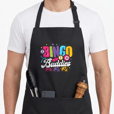 Buddy bingo aprons - Colorful BINGO Buddies text with numbered balls, hearts, stars, and bingo cards — shown on a black cotton canvas apron against a white background