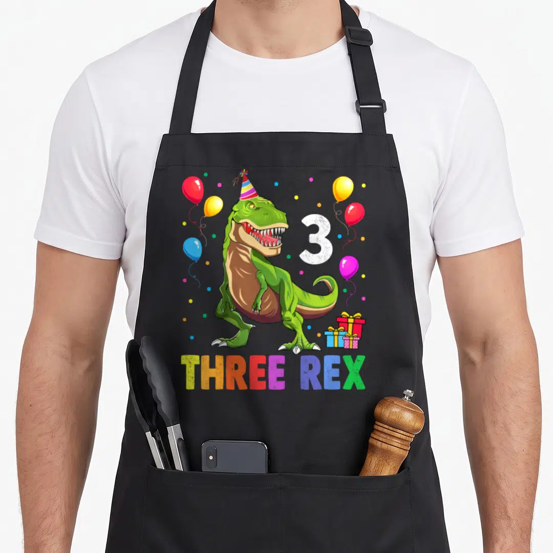 Third t-rex apron - Green T-Rex party hat THREE REX text balloons confetti gifts number 3 — shown on a black cotton canvas apron against a white background