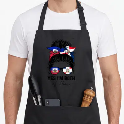 Haitian dominica apron - Woman head with Haitian Dominican flag glasses bun and Yes Im Both text — shown on a black cotton canvas apron against a white background