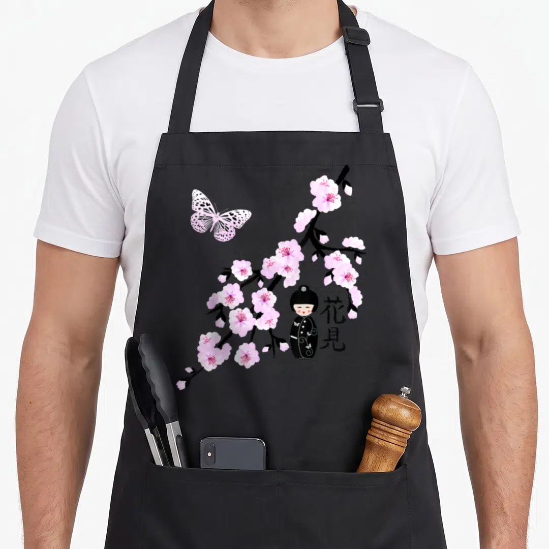 Character cherry apron - Pink cherry blossoms, black kokeshi doll, butterfly, and vertical kanji text on grey background — shown on a black cotton canvas apron against a white background