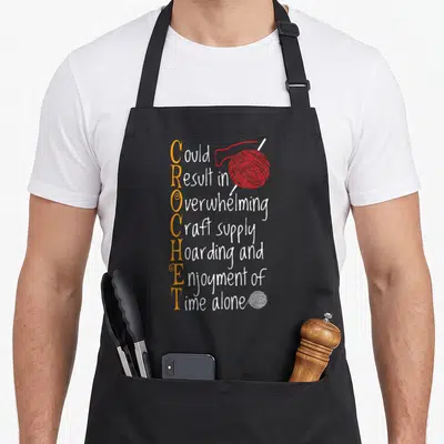 Sew crochet apron - Vertical orange CROCHET text with red and white yarn balls and stacked white sentence — shown on a black cotton canvas apron against a white background