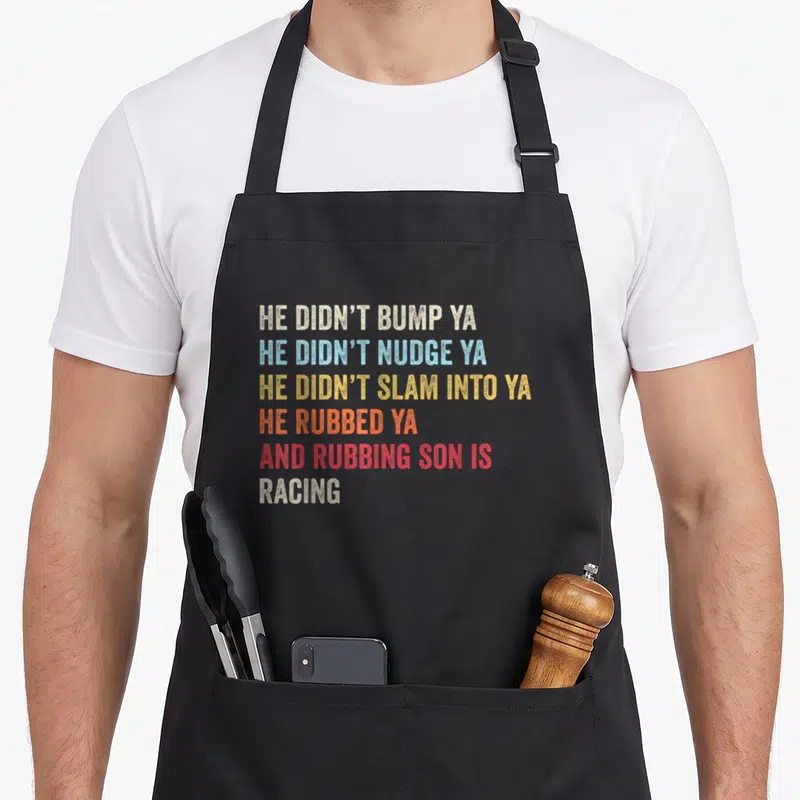 Car racing track apron - Stacked colorful text reading HE DIDN'T BUMP YA, HE DIDN'T NUDGE YA, HE DIDN'T SLAM INTO YA — shown on a black cotton canvas apron against a white background