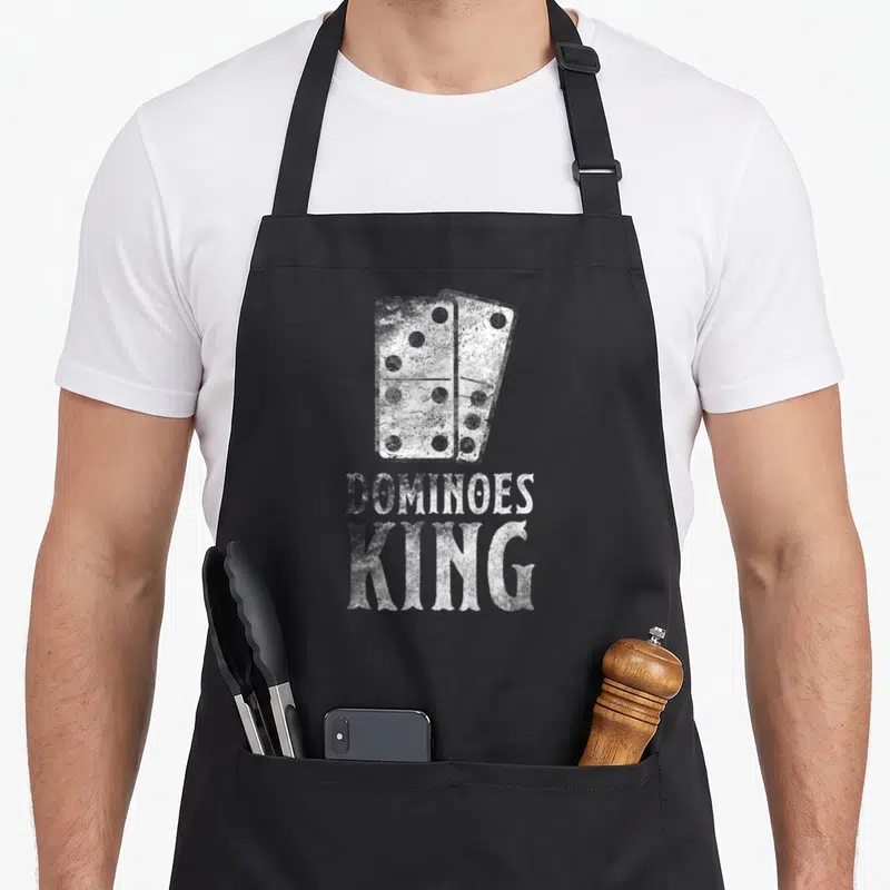 Domino king apron - Two white domino tiles overlapping above distressed DOMINOES KING text — shown on a black cotton canvas apron against a white background