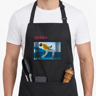 Break queen apron - Stacked QUEEN text above striped photo of singer in yellow leaning back with microphone — shown on a black cotton canvas apron against a white background