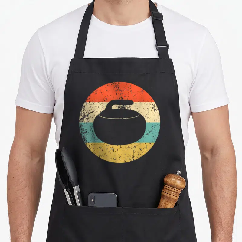Funny curling stone apron - Grey curling stone silhouette over four horizontal stripes in red, cream, teal, and yellow — shown on a black cotton canvas apron against a white background