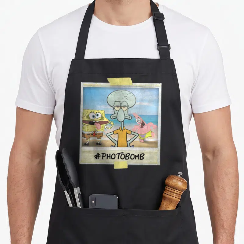 Squidward general apron - Squidward photobomb polaroid with hashtag text and tape details in cartoon style — shown on a black cotton canvas apron against a white background