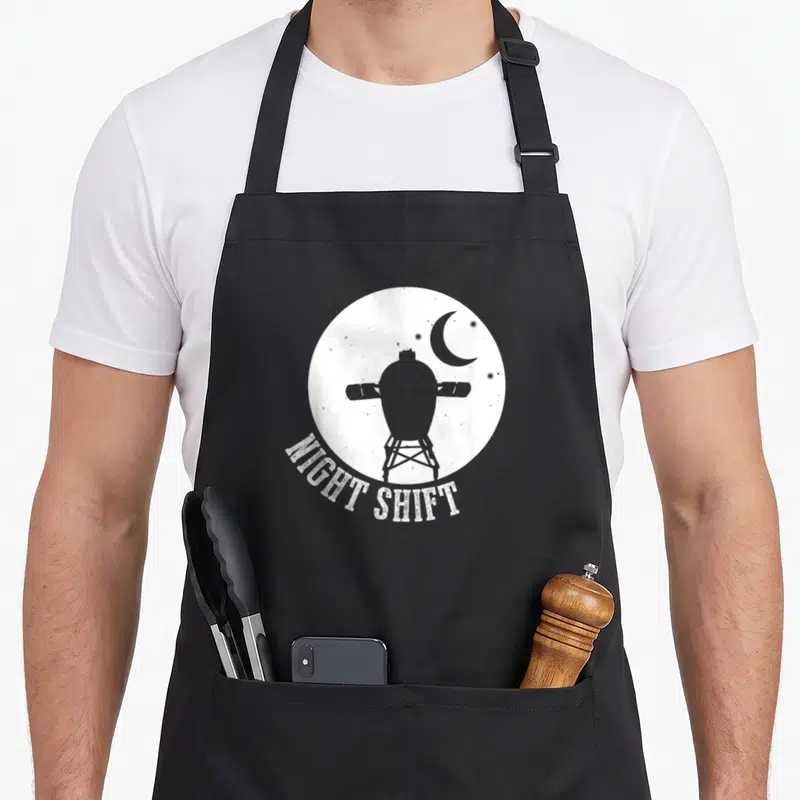 Night egg apron - Grey space capsule silhouette in white moon with arched NIGHT SHIFT text — shown on a black cotton canvas apron against a white background