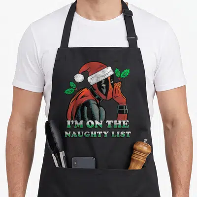 Secret santa apron - Distressed Deadpool in Santa hat flanked by holly leaves above two lines of text reading I'M — shown on a black cotton canvas apron against a white background