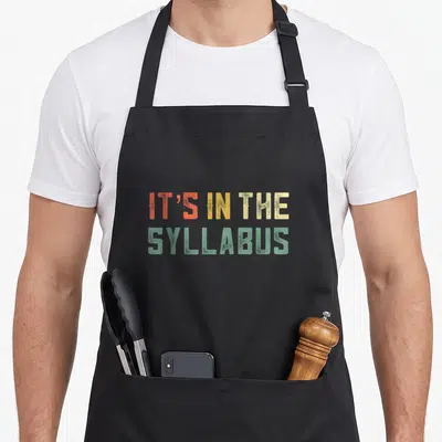 Tenure teacher apron - IT'S IN THE SYLLABUS text in orange yellow beige and green block letters — shown on a black cotton canvas apron against a white background