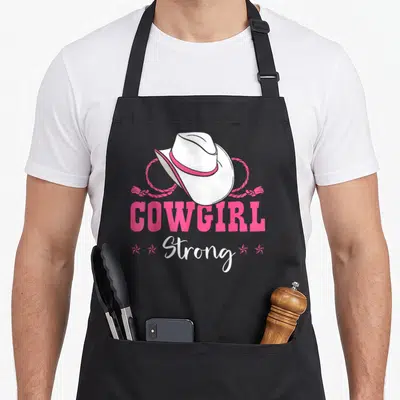 Barrel cowgirl aprons - White cowboy hat with pink band and rope above pink COWGIRL and white Strong text with four — shown on a black cotton canvas apron against a white background