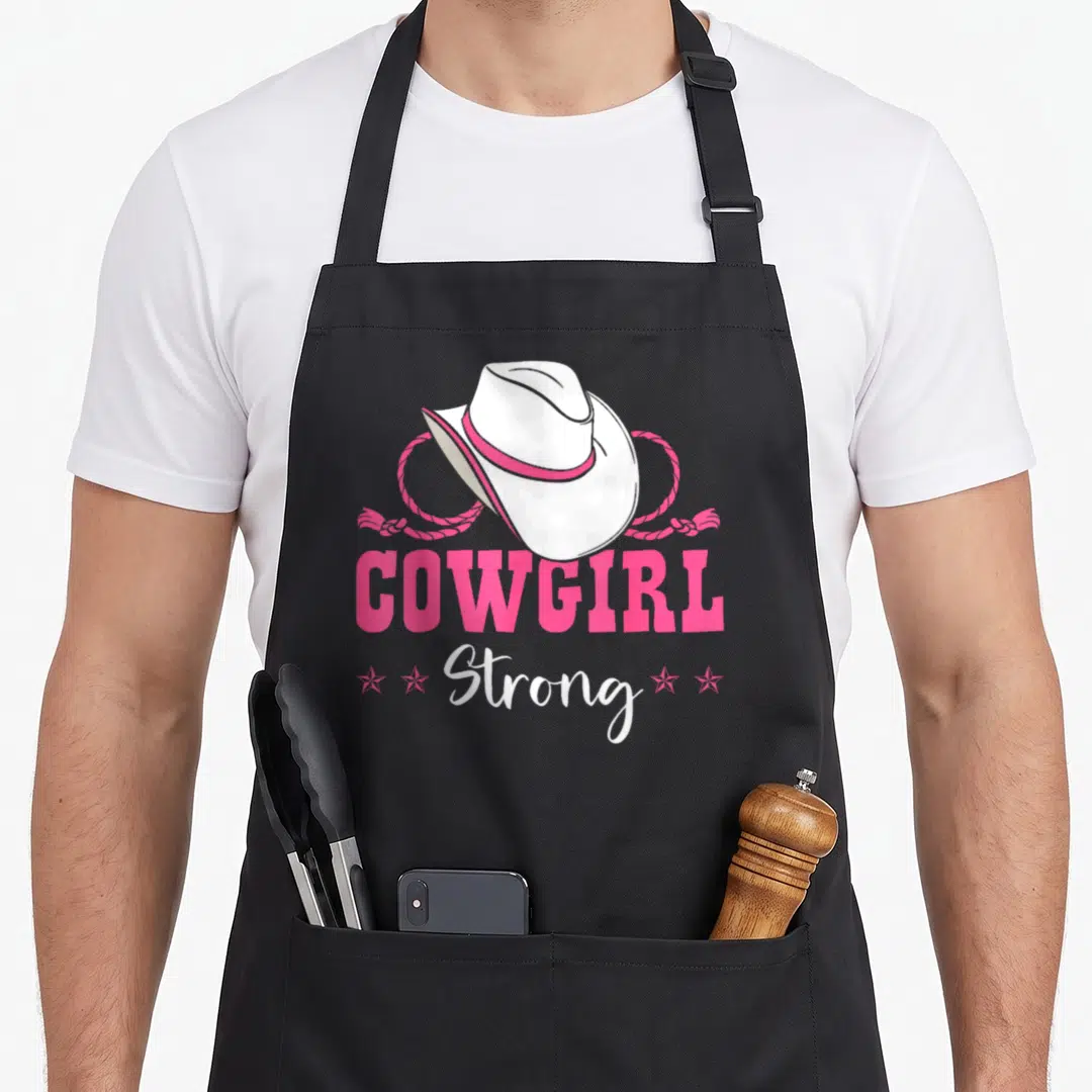 Barrel cowgirl aprons - White cowboy hat with pink band and rope above pink COWGIRL and white Strong text with four — shown on a black cotton canvas apron against a white background