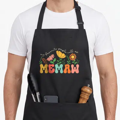 Cute call mother's day aprons - Retro MEMAW text with yellow red blue orange pink flowers and leaves — shown on a black cotton canvas apron against a white background