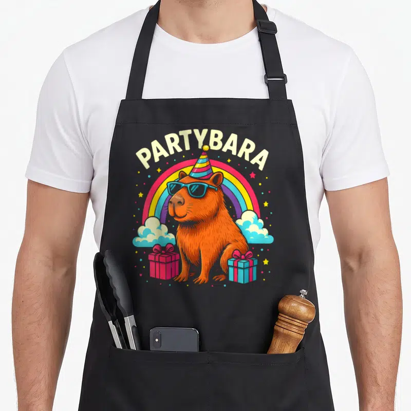 Partybara birthday apron - Cartoon orange capybara in sunglasses and party hat with rainbow and gifts under PARTYBARA — shown on a black cotton canvas apron against a white background