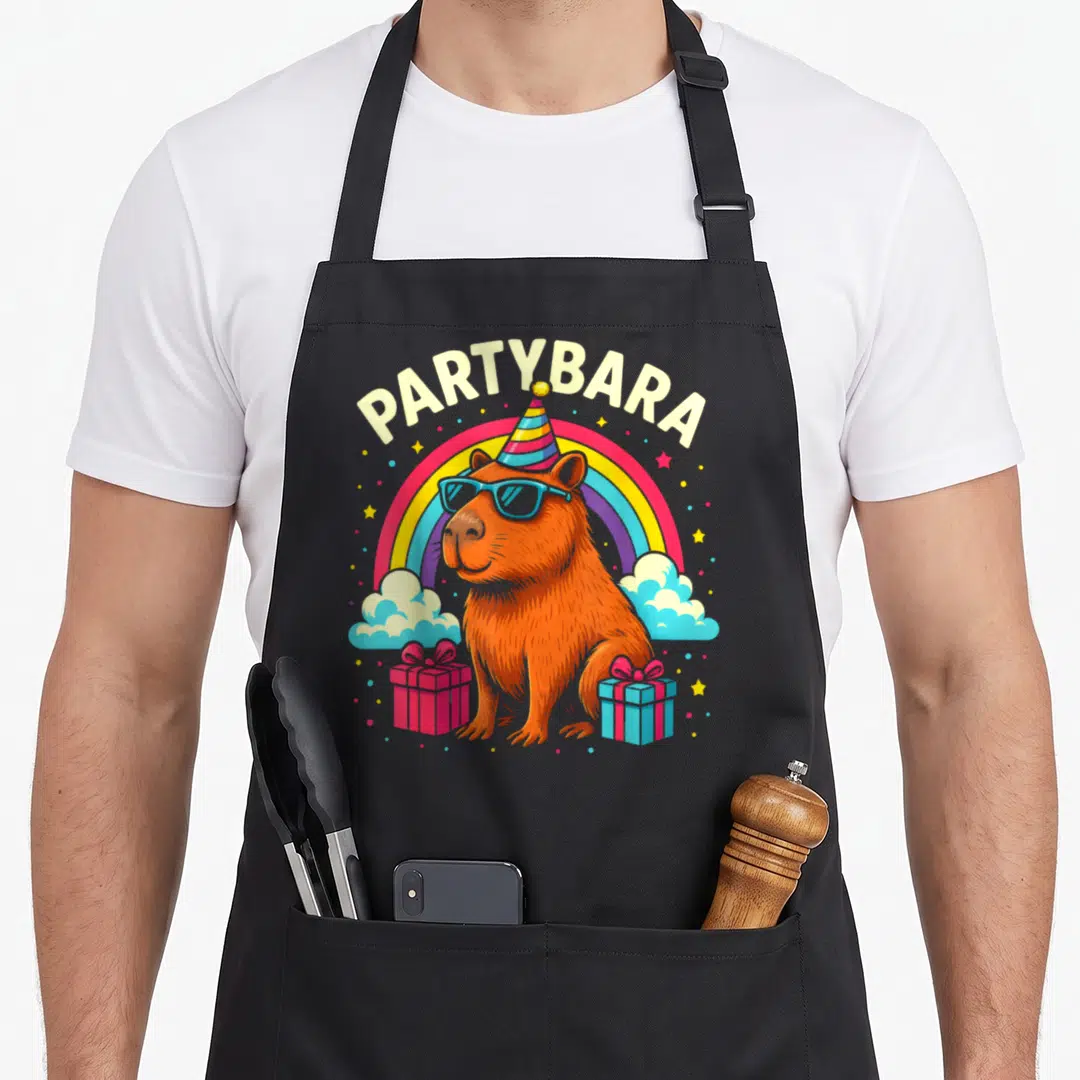 Partybara birthday apron - Cartoon orange capybara in sunglasses and party hat with rainbow and gifts under PARTYBARA — shown on a black cotton canvas apron against a white background