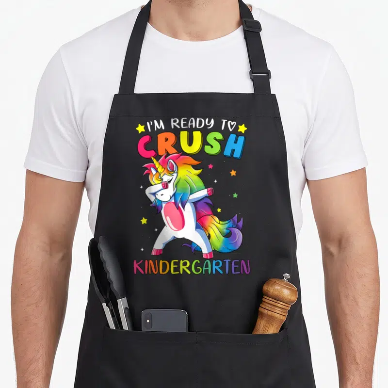 Crush unicorn aprons - Dabbing rainbow unicorn with I'M READY TO CRUSH KINDERGARTEN text and yellow stars — shown on a black cotton canvas apron against a white background