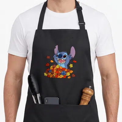 Autumn stitch apron - Blue cartoon alien emerging from a pile of orange, yellow, and red autumn leaves — shown on a black cotton canvas apron against a white background