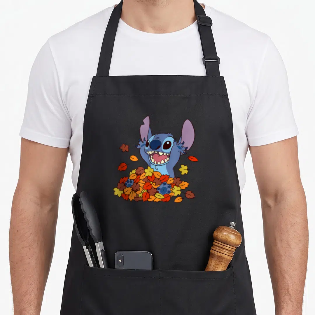 Autumn stitch apron - Blue cartoon alien emerging from a pile of orange, yellow, and red autumn leaves — shown on a black cotton canvas apron against a white background