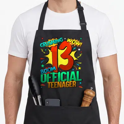 13th comic birthday apron - Red number 13 over yellow burst with CRUSHING WOW BOOM OFFICIAL TEENAGER text in comic style — shown on a black cotton canvas apron against a white background