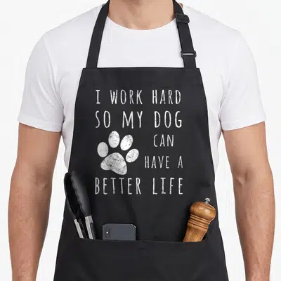 Hard better dog apron - White distressed paw print next to stacked text reading I work hard so my dog can have a better — shown on a black cotton canvas apron against a white background