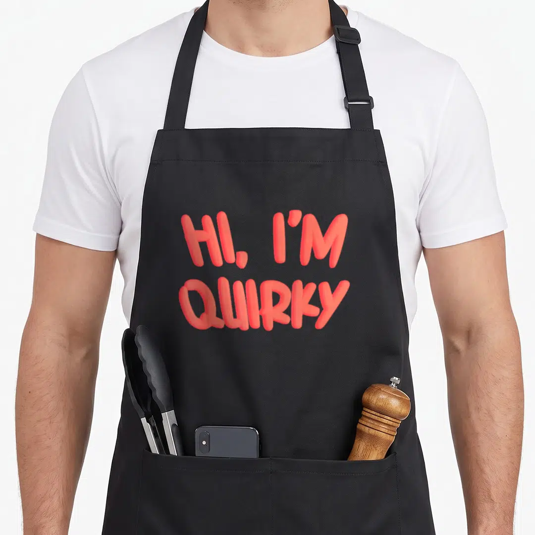 Funny quirky apron - Coral 3D style text reading HI I'M QUIRKY stacked in two lines on black fabric — shown on a black cotton canvas apron against a white background
