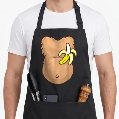 Monkey halloween apron - Cartoon hairy tan torso with a yellow peeled banana on the right side — shown on a black cotton canvas apron against a white background