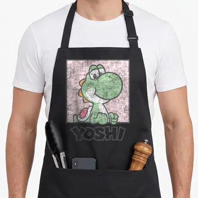 Simple pastel pink apron - Green cartoon Yoshi inside grey square frame on pink background with YOSHI text below — shown on a black cotton canvas apron against a white background