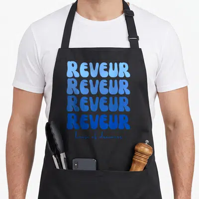 Dreamer teacher apron - Four stacked REVEUR words in blue gradient with house of dreamers script below — shown on a black cotton canvas apron against a white background