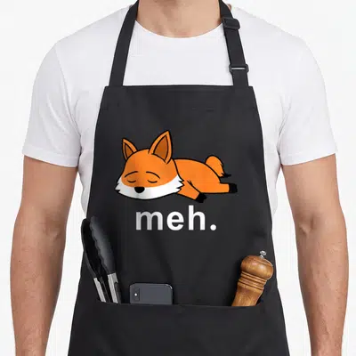 Fox meme apron - Cartoon orange fox lying down above white meh text on black fabric — shown on a black cotton canvas apron against a white background
