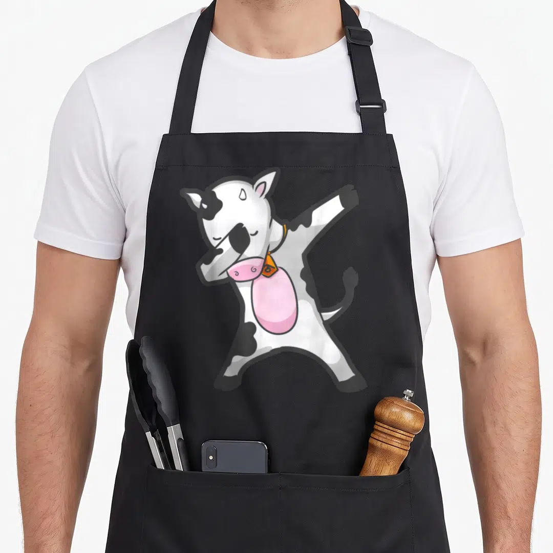 Dabbing cow apron - Cartoon cow dabbing with pink snout and orange bell — shown on a black cotton canvas apron against a white background