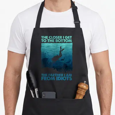 Scuba themed aprons - Diver silhouette in teal and blue water with text about getting farther from idiots at the bottom — shown on a black cotton canvas apron against a white background